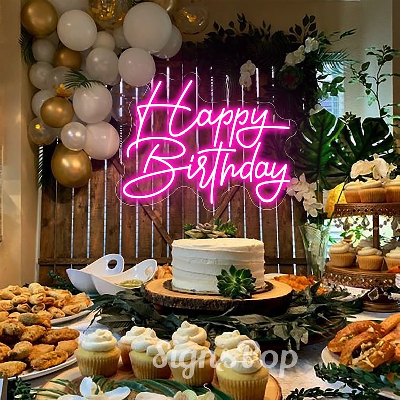 May include: Pink neon sign with the words "Happy Birthday" on a wooden background with a birthday cake and cupcakes in front of it.