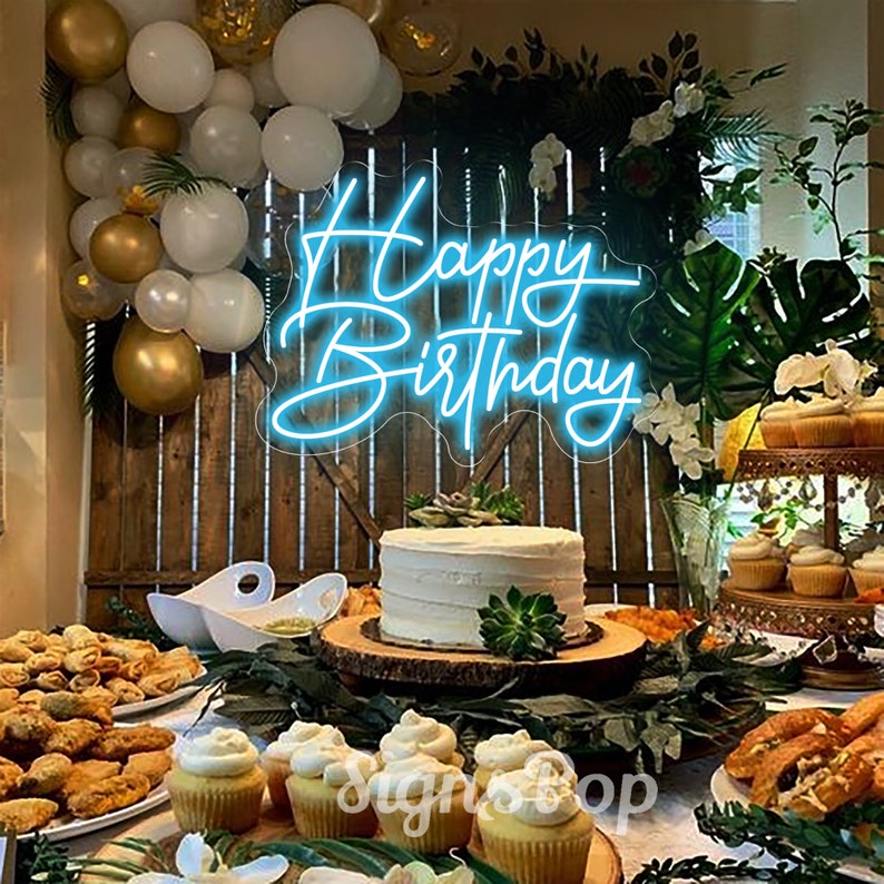May include: A blue neon sign with the words "Happy Birthday" on a wooden background. The sign is surrounded by balloons and greenery. There is a cake and cupcakes on a table in front of the sign.