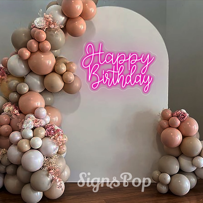 May include: A white backdrop with a pink neon sign that reads "Happy Birthday". The backdrop is decorated with a balloon arch made of beige, peach, and white balloons.