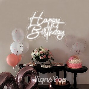 May include: White neon sign with the words "Happy Birthday" on a white wall. The sign is surrounded by balloons and a table with a cake and other decorations.