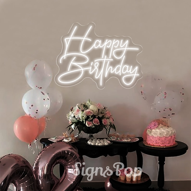 May include: White neon sign with the words "Happy Birthday" on a white wall. The sign is surrounded by balloons and a table with a cake and other decorations.