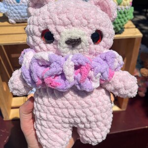 May include: A light pink crocheted bear plush toy with a soft, textured yarn finish. The bear features large, dark, glossy eyes, a small brown nose, and a ruffled collar made of variegated purple, pink, and white yarn around its neck.