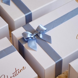 May include: White gift boxes tied with pale blue satin ribbons and bows. Some boxes feature names in gold script. The boxes are arranged on a textured, light-coloured surface, creating a visually appealing presentation.