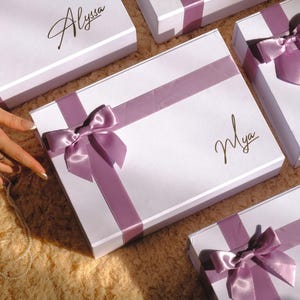 May include: White gift boxes with lavender satin ribbons and bows. Some boxes have names written in gold script. The boxes are arranged on a textured surface. The overall theme is gift-giving or special occasions.
