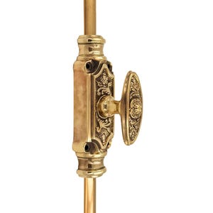 Könnte beinhalten: Ein Fensterriegel aus Messing mit einem verzierten floralen Design. Das Schloss hat eine rechteckige Basis mit detaillierten Schnitzereien und einen ovalen Griff. Die Hardware ist auf einer Messingstange montiert.