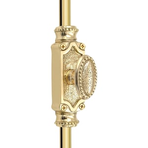 Puede incluir: Un cierre de ventana dorado con un asa ovalada y detalles decorativos. La cerradura está montada sobre una varilla dorada y presenta una placa trasera rectangular con bordes ornamentados y cabezas de tornillo en forma de cruz.