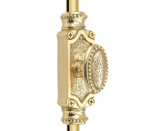 May include: A gold-colored window sash lock with an oval-shaped handle and decorative detailing. The lock is mounted on a gold-colored rod and features a rectangular backplate with ornate edges and cross-shaped screw heads.