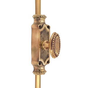 May include: A brass window sash lock featuring an oval knob and a rectangular backplate. The knob has a textured surface, and the backplate has an ornate design. The lock is mounted on a brass rod, with a vintage aesthetic.