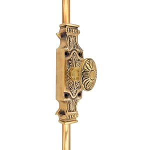 May include: A brass door bolt with an ornate design. The bolt features a rectangular plate with floral carvings and a round knob. The bolt is mounted on a vertical rod, and the overall design is antique.