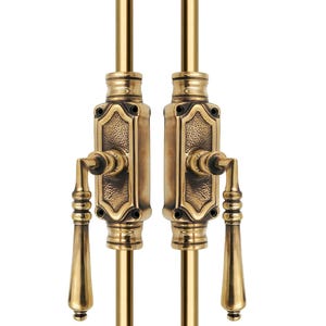 May include: Two antique brass window sash lifts. Each lift features a rectangular backplate with an ornate design, a lever handle, and a vertical rod. The brass has a warm, aged finish, suggesting vintage hardware.