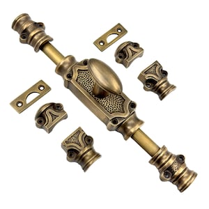 Puede incluir: Un juego de cerrojo de puerta de bronce de estilo antiguo. El juego incluye un cerrojo central largo con un asa ovalada, dos placas de cierre y cuatro piezas de montaje decorativas. El metal tiene un acabado cálido y envejecido, con detalles intrincados.