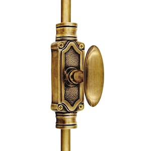 Könnte beinhalten: Nahaufnahme eines Fensterverschlusses aus Messing. Der Verschluss hat einen ovalen Griff und einen rechteckigen Körper mit dekorativen Details. Die Hardware hat eine warme, antike Goldfarbe und ist auf einer vertikalen Stange montiert.