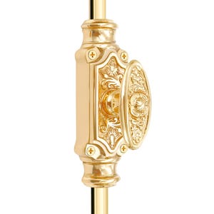 Puede incluir: Un pestillo de ventana dorado y ornamentado. La cerradura presenta un asa ovalada y una base rectangular, ambas con diseños detallados de flores y volutas. La ferretería está montada sobre una varilla dorada.