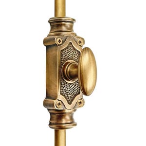 May include: Close-up of a brass-colored door latch. The latch features an oval handle and a rectangular plate with a textured pattern and decorative screws. The hardware is mounted on a vertical rod, suggesting a door or window fixture.