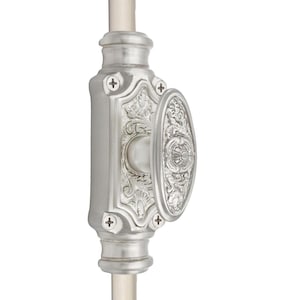 Può includere: Un chiavistello per porta argentato e ornato, con una maniglia ovale e motivi floreali decorativi. Il chiavistello è montato su un'asta verticale e presenta teste di viti a forma di croce.