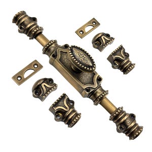 Puede incluir: Un cerrojo de puerta de bronce antiguo con un asa central ovalada y piezas decorativas en los extremos. El cerrojo va acompañado de varias placas de montaje y detalles decorativos a juego, todo en un estilo vintage.