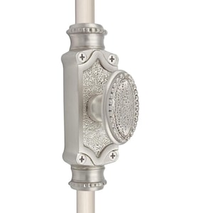 Puede incluir: Un pestillo de ventana plateado con un pomo ovalado. La cerradura tiene una placa trasera rectangular con detalles decorativos y está sujeta a una varilla blanca. El pomo y la placa trasera tienen una superficie texturizada.