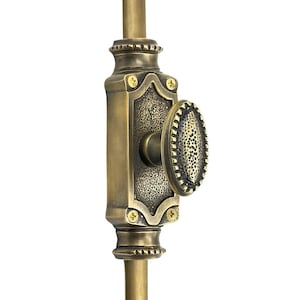 Puede incluir: Primer plano de un tirador de puerta de latón antiguo con un pomo de forma ovalada. El tirador tiene una superficie texturizada y detalles decorativos, incluyendo pequeños tornillos y una varilla vertical.