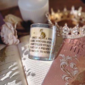 May include: A clear glass candle with a gold fox illustration and text that reads "ONCE UPON A TIME A GIRL WHO BELIEVED FAIRYTALES STOLE THE HEART OF A PRINCE WHO SWORN TO NEVER". The candle sits on a book with a gold crown in the background.