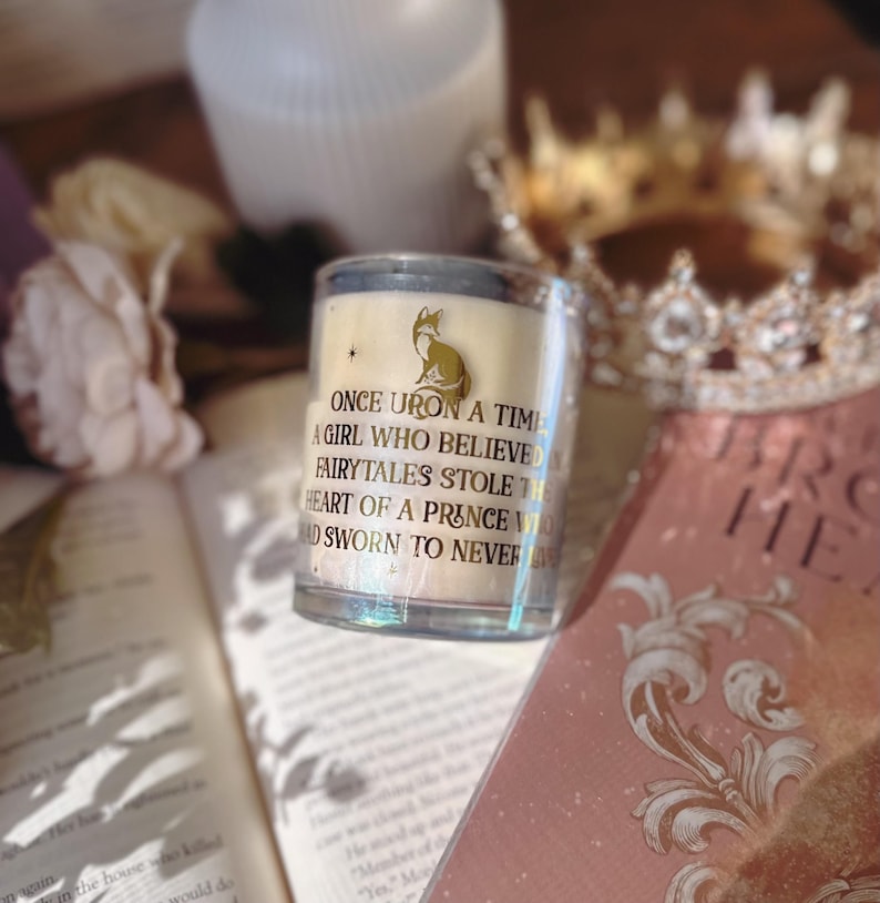 May include: A clear glass candle with a gold fox illustration and text that reads "ONCE UPON A TIME A GIRL WHO BELIEVED FAIRYTALES STOLE THE HEART OF A PRINCE WHO SWORN TO NEVER". The candle sits on a book with a gold crown in the background.