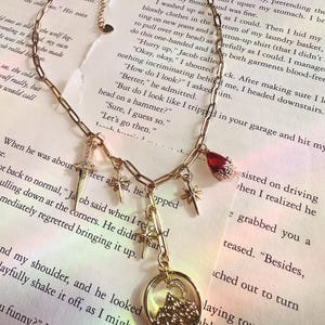 May include: Gold-toned necklace with multiple charms, including a cross, stars, a red gem, and a circular pendant with a mountain and moon design. The chain is on a background of printed text.