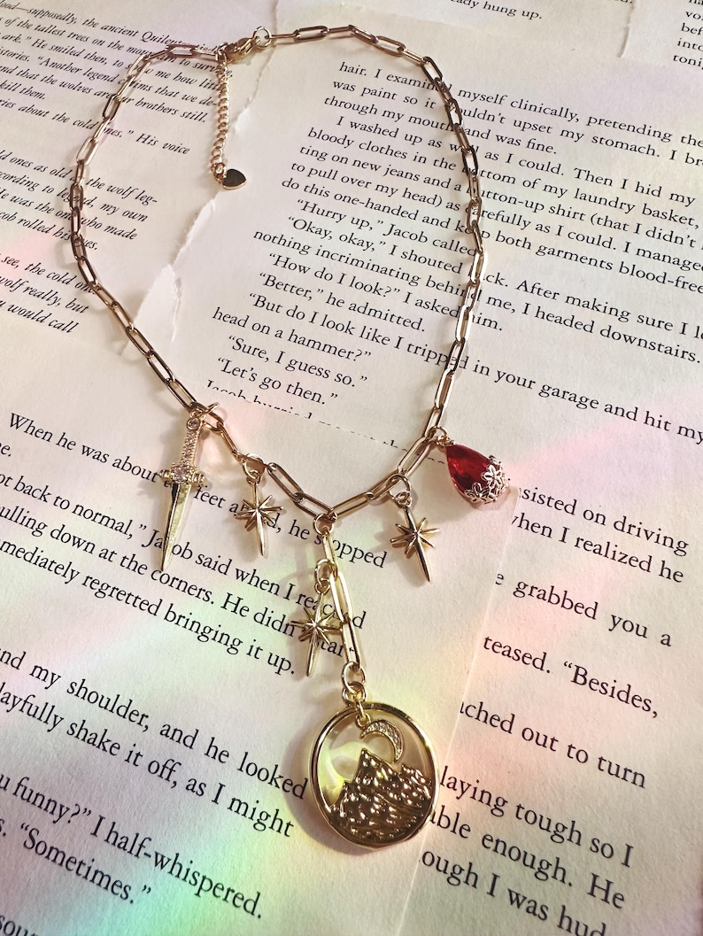 May include: Gold-toned necklace with multiple charms, including a cross, stars, a red gem, and a circular pendant with a mountain and moon design. The chain is on a background of printed text.