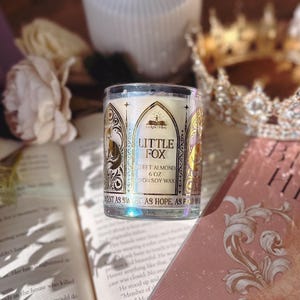 May include: A Little Fox Sweet Almond soy wax candle in a clear glass jar. The label features gold foil accents and the text "Little Fox" and "Sweet Almond". The candle is 6 oz. and is surrounded by books and a decorative crown.