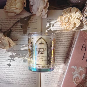May include: A clear glass candle jar with the text "LITTLE FOX SWEET ALMOND 6 OZ 100% SOY WAX" sits on an open book. The candle has a gold and white label with an arched design. The background includes flowers and a book.
