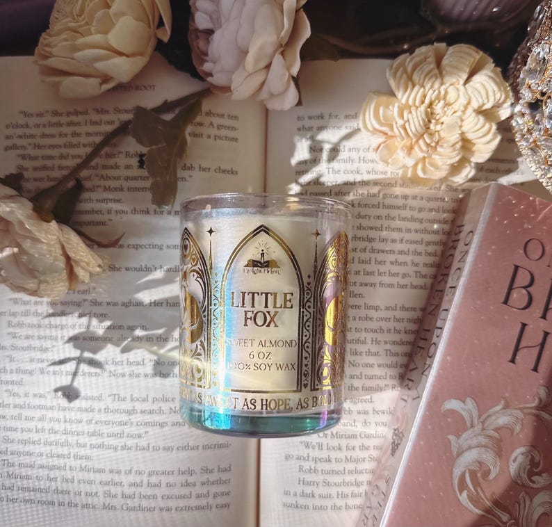 May include: A clear glass candle jar with the text "LITTLE FOX SWEET ALMOND 6 OZ 100% SOY WAX" sits on an open book. The candle has a gold and white label with an arched design. The background includes flowers and a book.