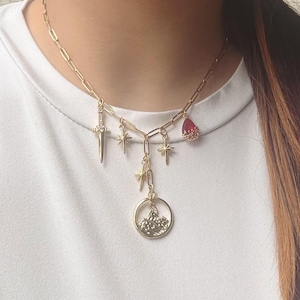 May include: Gold-toned necklace with multiple charms. The charms include a dagger, stars, a teardrop-shaped red gem, and a circular pendant with a mountain scene. The necklace is worn against a white background.