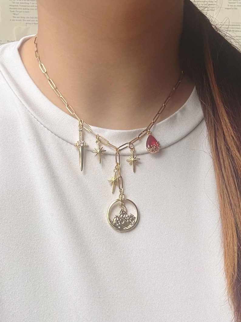 May include: Gold-toned necklace with multiple charms. The charms include a dagger, stars, a teardrop-shaped red gem, and a circular pendant with a mountain scene. The necklace is worn against a white background.
