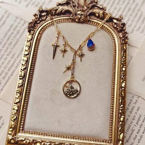Peut inclure: Un collier doré orné de plusieurs breloques : une dague, des étoiles, une pierre bleue en forme de goutte et un pendentif rond avec une montagne. Le collier est présenté dans un cadre doré.