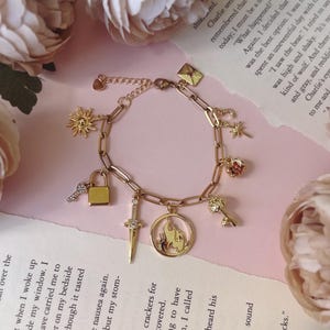 Puede incluir: Pulsera dorada con dijes variados: un sol, una llave, un candado, una espada, una estrella, una rosa y un colgante circular. La pulsera se muestra sobre una página de libro con flores rosas.