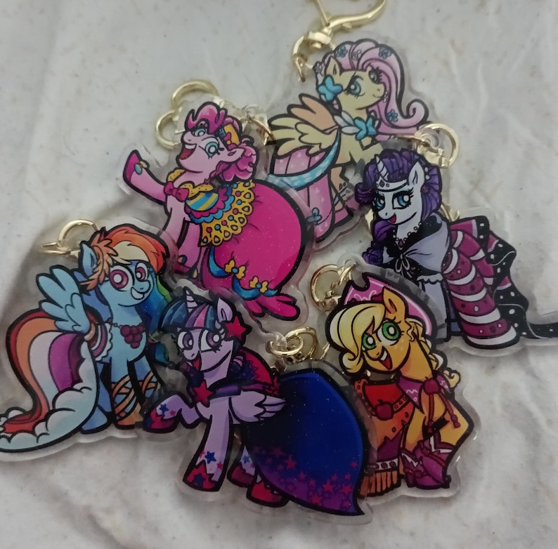 May include: A set of six colourful cartoon pony keychains. The keychains feature different pony characters with various hairstyles and accessories. The ponies are depicted in a whimsical style with bright colours and bold outlines.