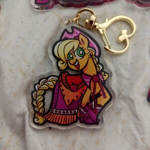 May include: A keychain featuring a cartoon character with blonde hair, wearing a pink and orange cowboy outfit. The character has green eyes and a bright smile. The keychain is made of clear acrylic and has a gold metal clasp.