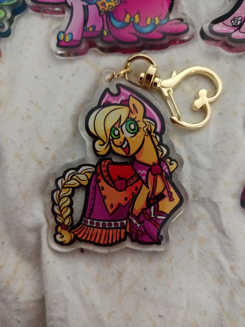 May include: A keychain featuring a cartoon character with blonde hair, wearing a pink and orange cowboy outfit. The character has green eyes and a bright smile. The keychain is made of clear acrylic and has a gold metal clasp.