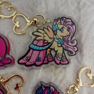 May include: A pink and yellow cartoon pony with a blue dress and wings, wearing a gold heart-shaped keychain. The pony has a pink mane and tail with blue flowers.