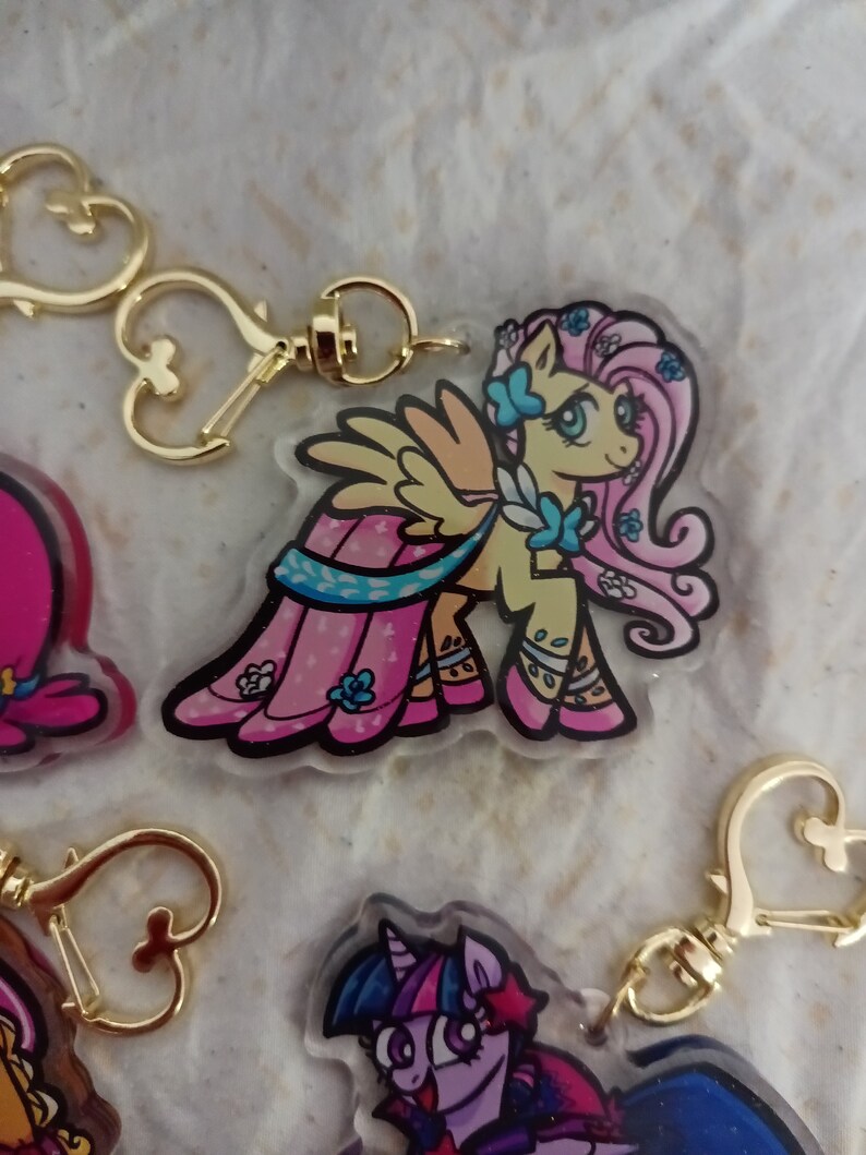 May include: A pink and yellow cartoon pony with a blue dress and wings, wearing a gold heart-shaped keychain. The pony has a pink mane and tail with blue flowers.