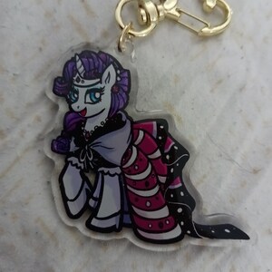 May include: A purple and white cartoon pony with a crown and a pink and white dress. The pony is holding a rose in its hoof. The pony is attached to a gold keychain.