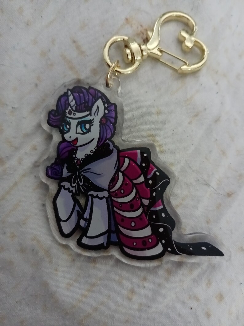 May include: A purple and white cartoon pony with a crown and a pink and white dress. The pony is holding a rose in its hoof. The pony is attached to a gold keychain.