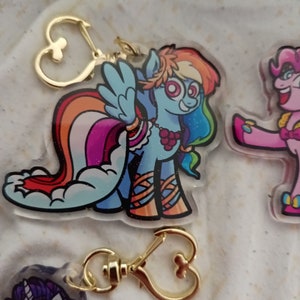 May include: A colourful cartoon character keychain featuring a rainbow pony with a pink, orange, yellow, green, blue, and purple mane and tail. The pony is wearing a red and white striped shirt and a necklace of red berries. The keychain has a gold heart-shaped clasp.