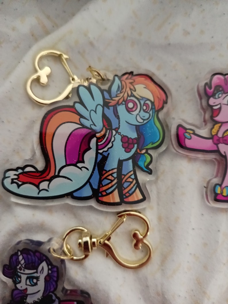 May include: A colourful cartoon character keychain featuring a rainbow pony with a pink, orange, yellow, green, blue, and purple mane and tail. The pony is wearing a red and white striped shirt and a necklace of red berries. The keychain has a gold heart-shaped clasp.