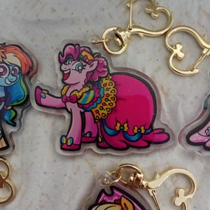 May include: Pinkie Pie from My Little Pony is wearing a pink and blue gown with gold accents. She is holding a bouquet of flowers and smiling. The charm is attached to a gold chain with a heart-shaped clasp.