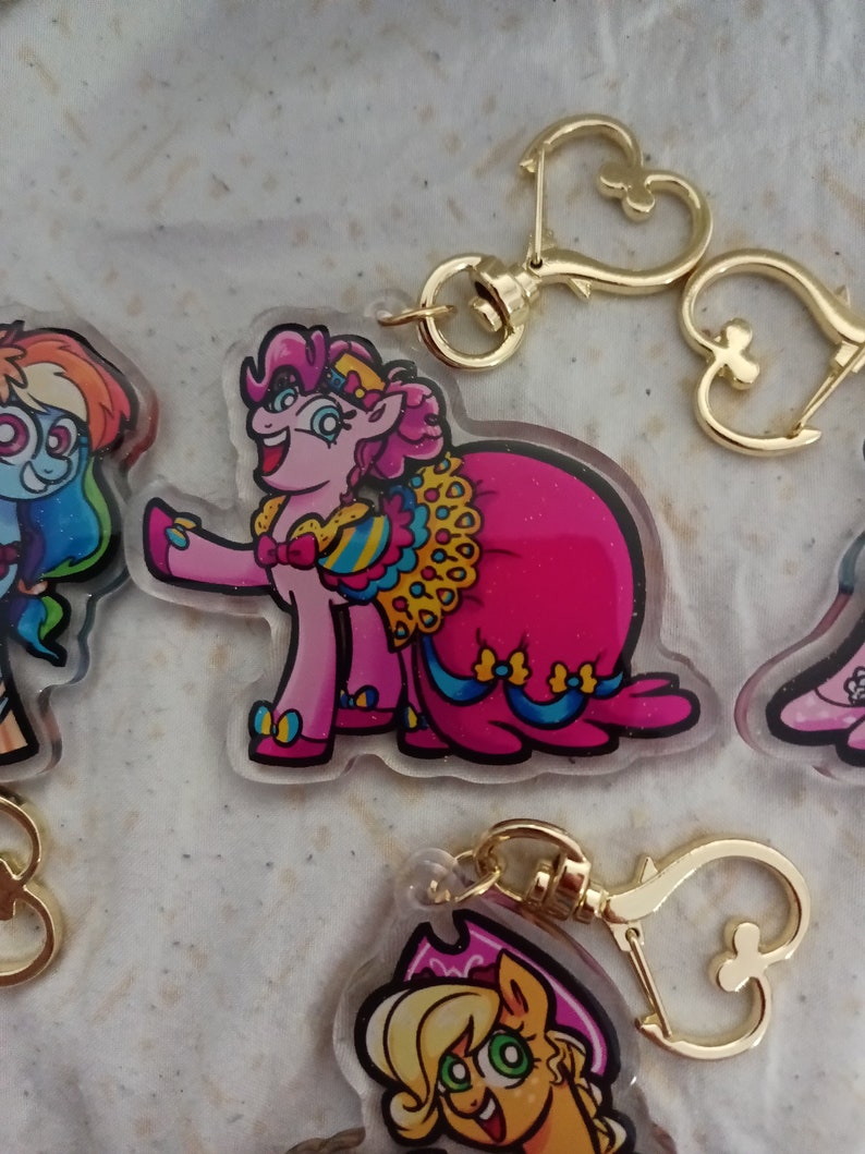 May include: Pinkie Pie from My Little Pony is wearing a pink and blue gown with gold accents. She is holding a bouquet of flowers and smiling. The charm is attached to a gold chain with a heart-shaped clasp.