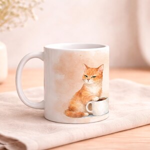 May include: White ceramic mug featuring a watercolor illustration of an orange tabby cat sitting next to a small white coffee cup. The cat has a grumpy expression. The background has a peach-colored watercolor wash.