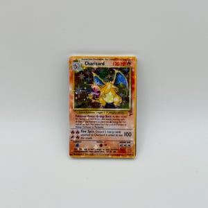 May include: A holographic Charizard trading card with 120 HP, featuring a fire-breathing dragon illustration, the Energy Burn Pokémon Power, and the Fire Spin attack. The card displays a star-patterned foil finish and detailed game text.