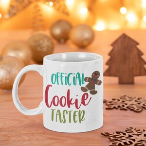May include: White ceramic mug with a gingerbread man graphic and the text "OFFICIAL Cookie TASTER" in colorful lettering. The mug is sitting on a wooden surface with a blurred background of golden lights and ornaments.