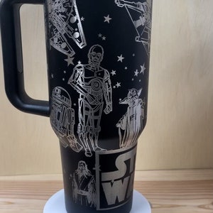 May include: Black travel mug with a silver Star Wars design featuring characters like Yoda, R2-D2, C-3PO, and a TIE fighter. The mug has a black handle and a silver lid.