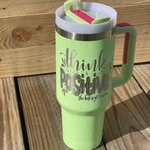以下が含まれることがあります： ピンクの取っ手と透明な蓋が付いた、薄い緑色のステンレス製のトラベルマグ。マグには、「Think Positive The best is yet to come」という銀色の文字が刻まれています。