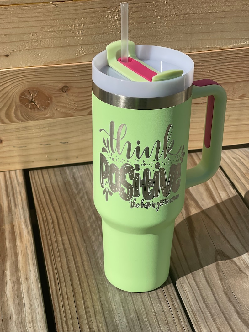 以下が含まれることがあります： ピンクの取っ手と透明な蓋が付いた、薄い緑色のステンレス製のトラベルマグ。マグには、「Think Positive The best is yet to come」という銀色の文字が刻まれています。